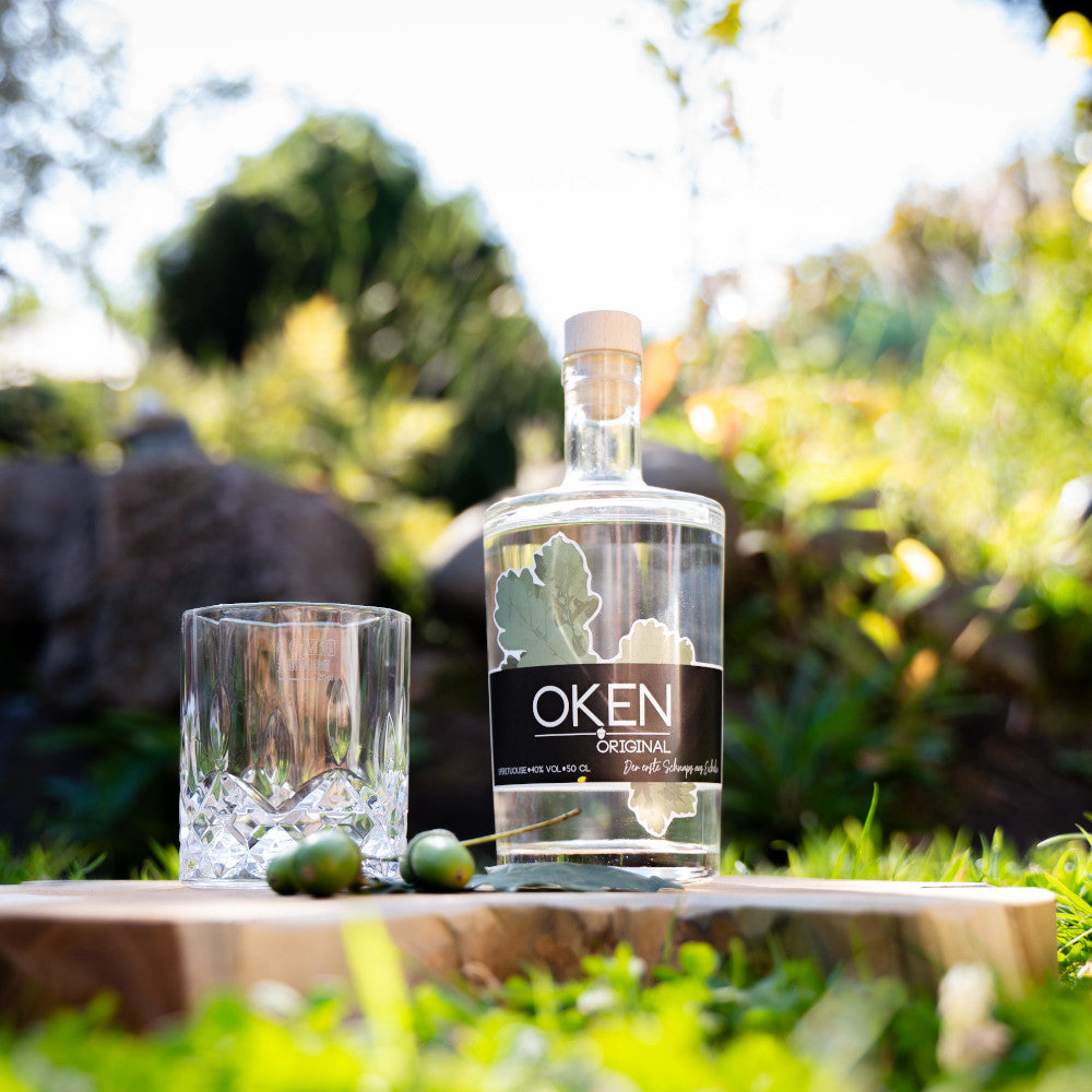 OKEN Original - Edelbrand aus Eicheln. Sehr mild, Aromen von Birne/Mirabelle und hellem Holz. Flasche OKEN im Garten mit Tumbler.